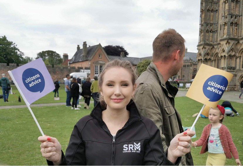 Rosie Carver holding two small flags with the Citizens Advice Mendip logo, standing outside Wells Cathedral.